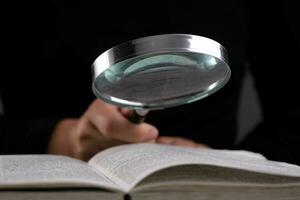 Close-up of a woman looking through a magnifying glass at a textbook. Magnifying glass in hand and open book on table. Education and research concept. photo