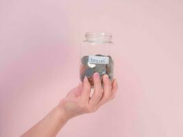 Close-up of young woman's hand holding glass jar with money  inside on pink background. Hand holding a money jar, travel, savings, education, donation. Finance plan concept. photo