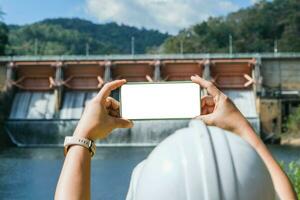 Environmental engineer using mobile phone to take picture with white screen against dam background. Water and ecology concept. photo