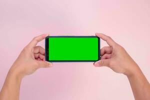 Human hand holding mobile smartphone with green screen in horizontal position isolated on pink background. clipping path photo