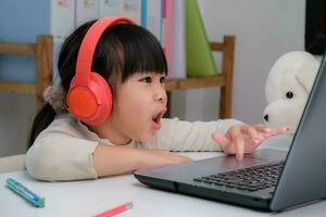 Cute elementary school girl wearing headphones and using a laptop computer. Happy Asian kids study online interactively with laptop computer or homeschooling, listening to music or playing games. photo
