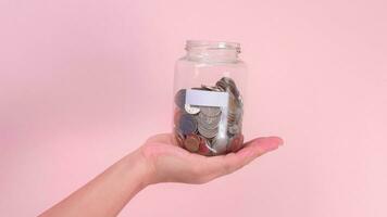 Close-up of hand holding glass jar with money  inside on pink background. Hand holding a money jar, travel, savings, education, donation. Finance plan concept. photo