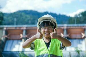 Portrait of a little girl engineer wearing a green vest and white helmet smiling happily on the background of the dam. Concepts of environmental engineering, renewable energy and love of nature. photo