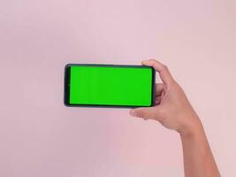 Human hand holding mobile smartphone with green screen in horizontal position isolated on pink background. clipping path photo