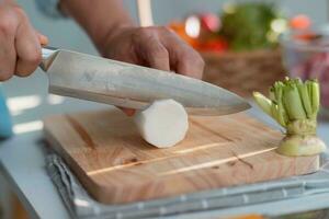 Cooking - Chef's hand cutting white radish on a chopping board in the kitchen. Preparing pork stock with vegetables broth in a pot. Homemade bouillon recipe. photo