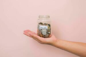 Close-up of young woman's hand holding glass jar with money coi inside on pink background. Hand holding a money jar, travel, savings, education, donation. Finance plan concept. photo