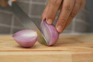 Female hand using knife to slice fresh red onion on chopping board, close-up. Chef chopping shallots using a sharp knife on a wooden chopping board. Preparing homemade food photo