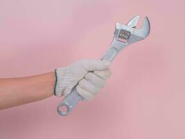 Hand in white knitted glove holding metal wrench isolated on pink background. Worker's hand in a glove holding a wrench. photo