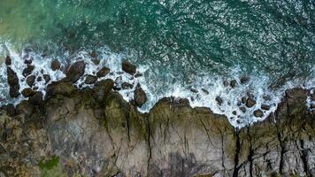 Aerial view of sea waves crashing on rocks cliff in the blue ocean. Top view of coastal rocks in Phuket ocean. Landscape view point of Laem Phromthep Cape in the morning. photo