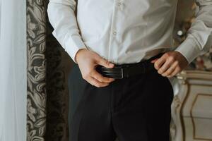 un joven hombre sujeta su cinturón en su pantalones en su habitación, de cerca foto. el novio es preparando para el Boda ceremonia foto