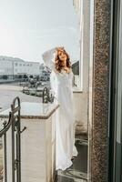 frio mujer en blanco atuendo sonriente en terraza. contento joven dama en elegante vendaje vestido poses en balcón y disfruta soleado clima foto