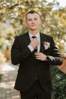 young groom fastens his tie looking into the lens outdoors. An adult man in a black suit and white shirt with a tie photo