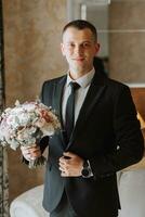 young man with a bouquet of flowers in a hotel room by the window, an adult man in a black suit and white shirt with a tie. The groom is preparing for the wedding ceremony. Close-up photo. photo