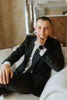 a young man is sitting in a chair in his room, near a window in a hotel room, a young businessman is wearing a black suit and a white shirt with a tie. The groom is preparing for the wedding ceremony. photo