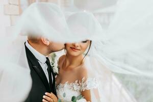 Stylish newlywed European couple. Smiling bride in a white dress. The groom, dressed in a classic black suit, white shirt, kisses the bride on the cheek under the veil. Wedding in nature photo
