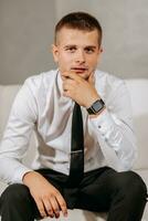 A mature man in a white shirt and black pants is sitting on a beige sofa in his room. Preparation for the wedding ceremony. Portrait of the groom. Free space photo
