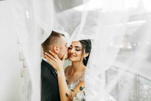 Stylish newlywed European couple. Smiling bride in a white dress. The groom, dressed in a classic black suit, white shirt, kisses the bride on the cheek under the veil. Wedding in nature photo