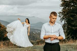 Beautiful bride and groom on the background of autumn mountains in front. A luxurious white dress flutters. The bridegroom is in the foreground. Wedding ceremony on top of the mountain. Free space. photo