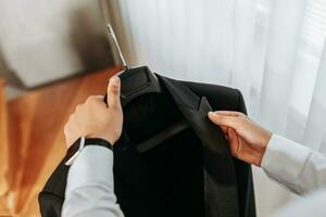 A man's hands hold a black jacket on a hanger by the window in his room. Preparation for the wedding ceremony. Close-up photo of hands