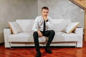 A mature man in a white shirt and black pants is sitting on a beige sofa in his room. Preparation for the wedding ceremony. Portrait of the groom. Free space photo