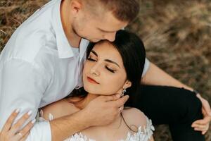 Groom and bride lie on dry grass and hug, woman in a white wedding dress. Beautiful autumn wedding photo. The groom aims at her forehead photo