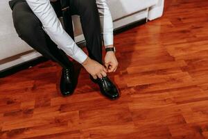 A mature man in a white shirt and black pants is putting on his shoes while sitting on the sofa in his room. Preparation for the wedding ceremony. Close-up photo. Free space photo