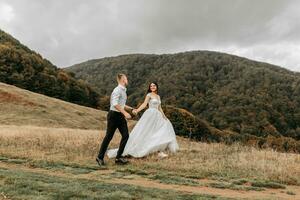 A romantic fairytale couple of newlyweds are running in a field at sunset, behind high mountains covered with trees. The bride in a white wedding dress, the groom in a white shirt and black pants photo