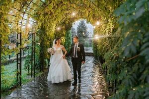 The bride and groom are walking in the evening park with a bouquet of flowers and greenery, greenery in nature. Romantic couple of newlyweds outdoors. Wedding ceremony in the botanical garden. photo