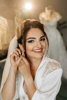 The morning of the bride in a white robe and an elegant hairstyle. Portrait of a girl fastening an earring, looking into the camera lens and smiling photo