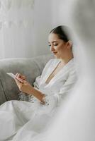 Portrait of a beautiful bride girl with stylish hairstyle and makeup in a white robe sitting on a sofa and reading from a white paper photo