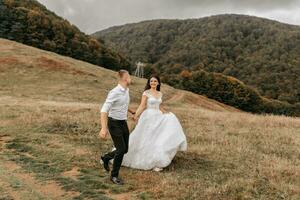 A romantic fairytale couple of newlyweds are running in a field at sunset, behind high mountains covered with trees. The bride in a white wedding dress, the groom in a white shirt and black pants photo