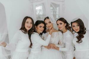 joven damas de honor en blanco seda vestidos abrazo y alegrarse en el de la novia habitación. hermosa mujer celebrando despedida de soltera fiesta en pie en habitación. foto