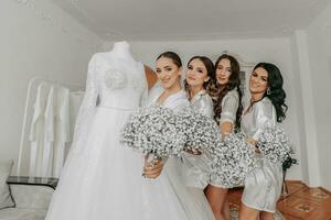 The bride's friends together with the bride hold bouquets of gypsophila flowers near the wedding dress. The bride and her fun friends celebrate the bachelorette party in matching dresses. photo