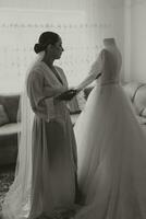 Stylish bride with wedding hairstyle and makeup in white robe standing near her wedding dress. Black and white photo