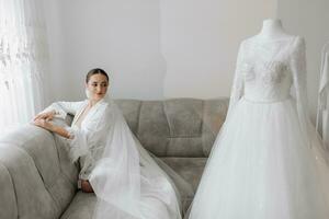 A wide-angle portrait of a beautiful bride with stylish hair and makeup in a white robe, sitting on a luxurious gray sofa and looking at her wedding dress. photo