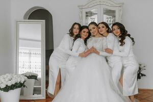 Young bridesmaids in white silk dresses hug and rejoice in the bride's room. Beautiful women celebrating bachelorette party standing in room. photo