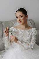 portrait of a beautiful bride with wedding hairstyle and makeup, sitting on a sofa in a wedding dress and holding a gypsophila flower in her hands, looking into the camera lens photo