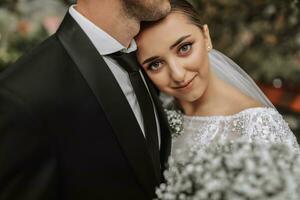 A stylish couple of European newlyweds. A smiling bride in a white dress lies on the groom's shoulder. The groom, dressed in a classic black suit, white shirt, . Wedding in nature photo