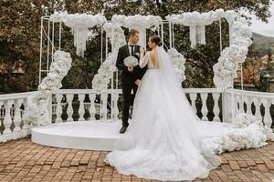 Stylish groom in a suit and cute brunette bride in a white dress in a park near a wedding arch decorated with white flowers. Wedding portrait of newlyweds. photo