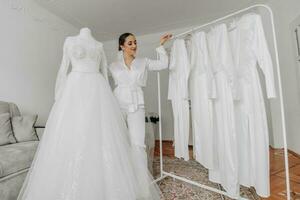 a beautiful bride poses in a white robe next to her wedding dress and her friends' dresses on a hanger photo