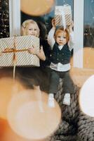 Merry Christmas and Happy Holidays. Cheerful girl and boy are sitting on the windowsill near the Christmas tree with Christmas presents. Children are having fun. Loving family with gifts in the room. photo