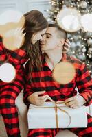 Young beautiful family couple in identical red pajamas near the Christmas tree rejoice and exchange Christmas gifts in their hands. New Year's holidays and gifts under the Christmas tree photo