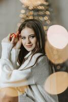 un hermosa y elegante joven niña en un blanco suéter poses en el Navidad luces en el antecedentes de el Navidad árbol foto