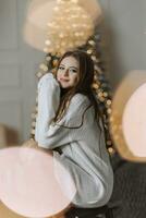 un hermosa y elegante joven niña en un blanco suéter poses en el Navidad luces en el antecedentes de el Navidad árbol foto
