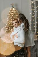 A beautiful and stylish young family in white sweaters hug and rejoice in the Christmas lights against the background of the Christmas tree. New year concept photo