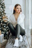 Beautiful and stylish young girl in white sweater posing sitting on windowsill in Christmas lights and Christmas tree with gifts photo