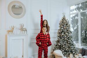 un hermosa joven niña en rojo pijama soportes en el antepecho y pone en un Navidad árbol. Navidad concepto. muchos regalos en papel envoltorios debajo el Navidad árbol foto