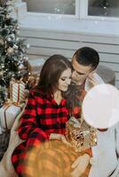 Young beautiful family couple in identical red pajamas near the Christmas tree, wrapped in a blanket, rejoice and exchange New Year's gifts. New Year's holidays and gifts under the Christmas tree photo