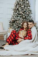 Young beautiful family couple in identical red pajamas near the Christmas tree, wrapped in a blanket, rejoice and exchange New Year's gifts. New Year's holidays and gifts under the Christmas tree photo