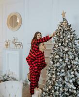 a beautiful young girl in red pajamas stands on the windowsill and puts on a Christmas tree. Christmas concept. Many gifts in paper wrappers under the Christmas tree photo